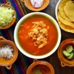 plato de pozole rojo sobre una mesa con mantel maxicano y al rededor del pozole sus complementos