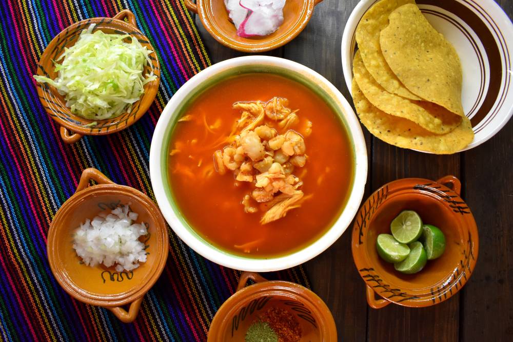 plato de pozole rojo sobre una mesa con mantel maxicano y al rededor del pozole sus complementos